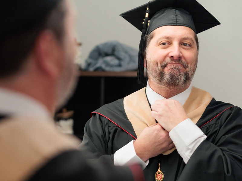 A man in a cap and gown adjusts his tie in the mirror