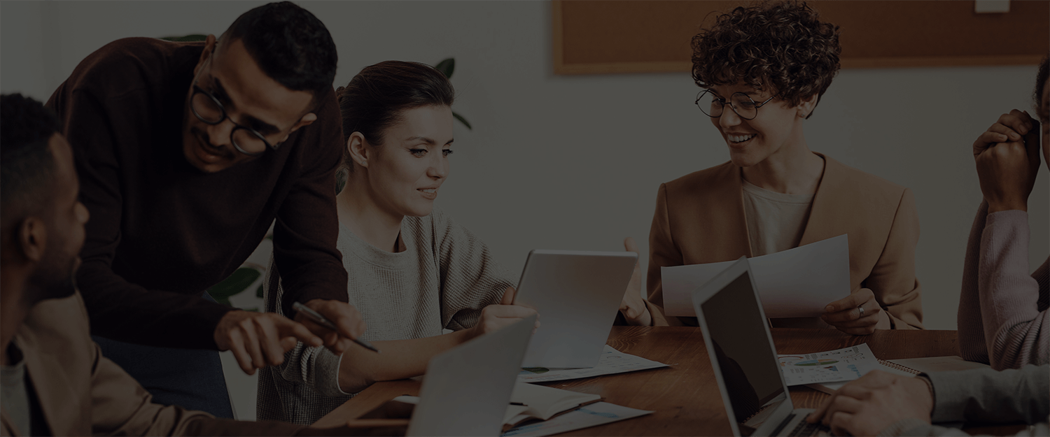 Six business professionals are sitting in a roundtable meeting talking amongst themselves while looking at tablets, laptops, and documents.
