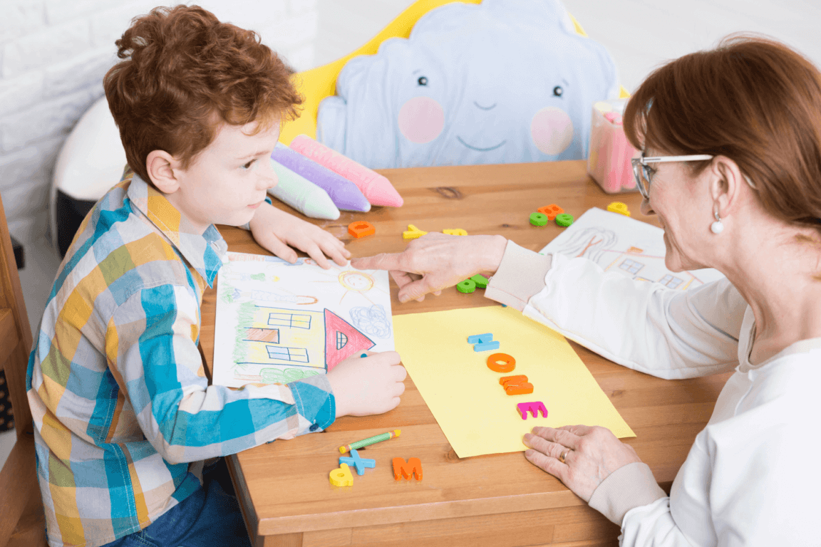 A teacher sits with a child at a wooden table. The child is holding a picture with a drawing of a house, while the teacher holds a yellow piece of paper that has "HOME" blocks on it.