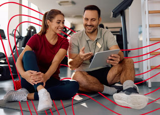 Health coach sitting on a gym floor reviewing fitness progress with a client on a tablet in a workout facility.