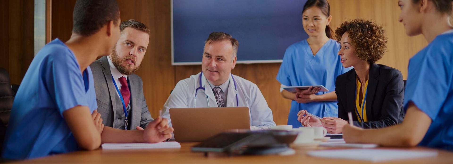 Six health professionals sit around a wooden table during a meeting.