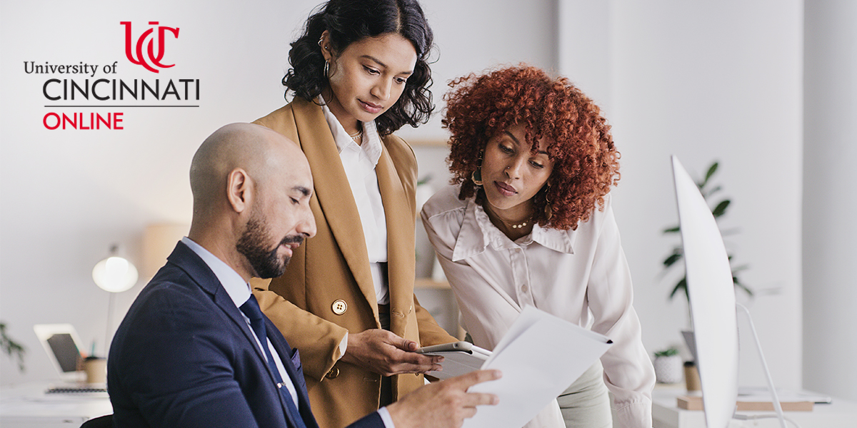Three business professionals observe a sheet of paper during a meeting. One person is seated while two are standing.