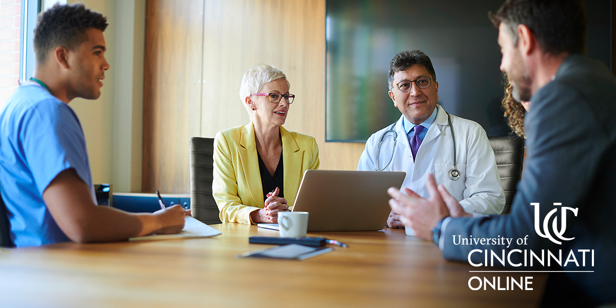 A group of health professionals sits at a meeting table speaking with each other. There are five people at the table.