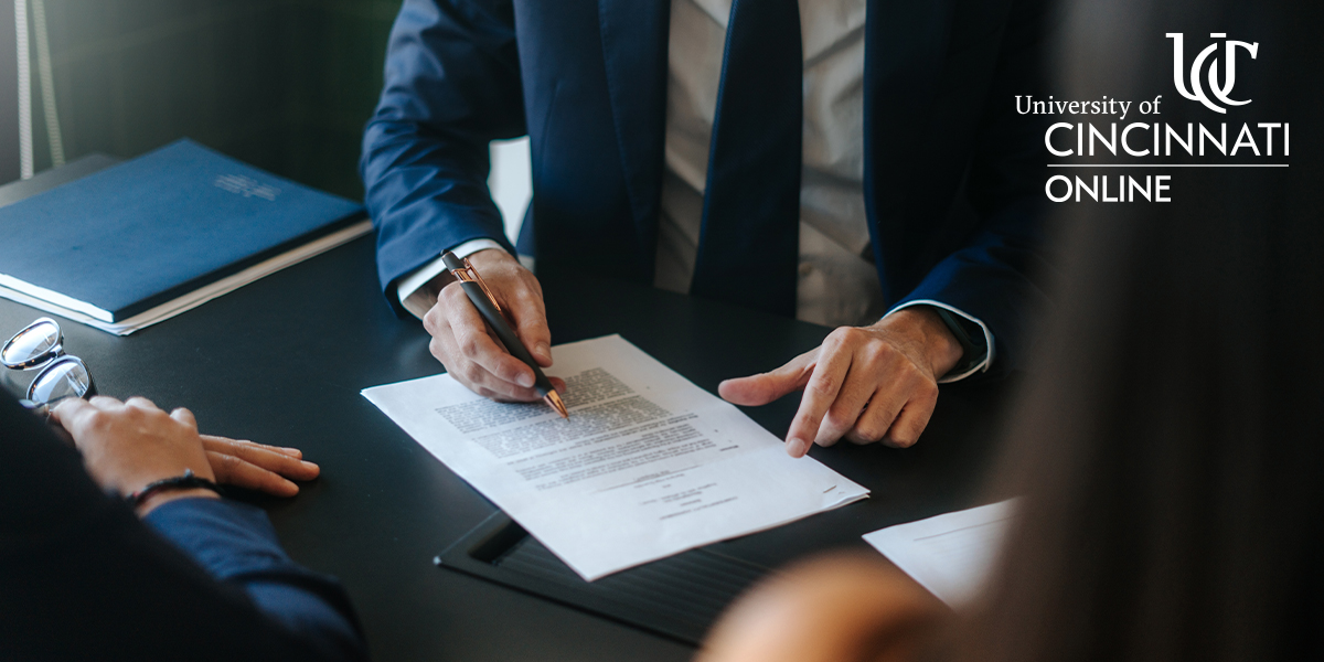 Three people in business attire sit at a table while one person goes over a legal document while holding a pen in their right hand. You can't see anyone's faces in the image.