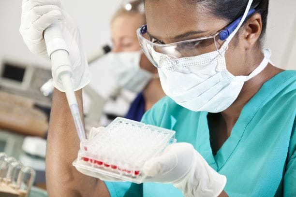 A lab scientist wearing a mask, safety glasses, and gloves handles a syringe and blood samples.