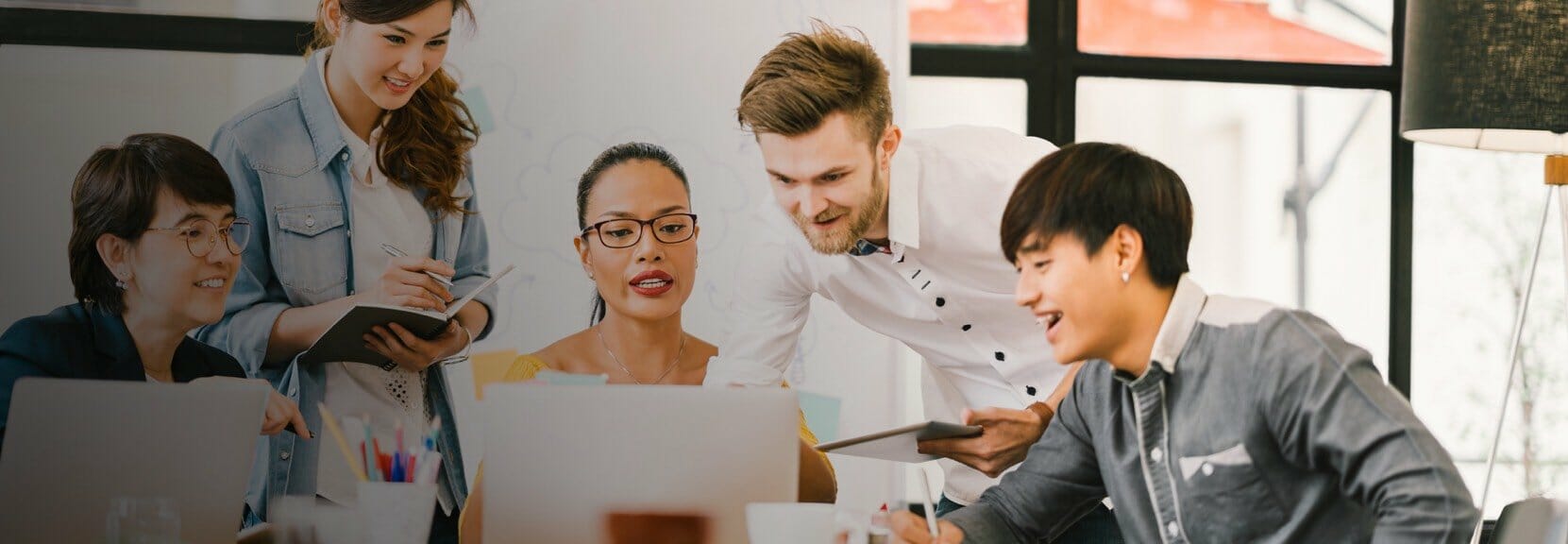 A woman in glasses sits at a table with an open laptop while leading a meeting as four colleagues takes notes and look over her shoulder.