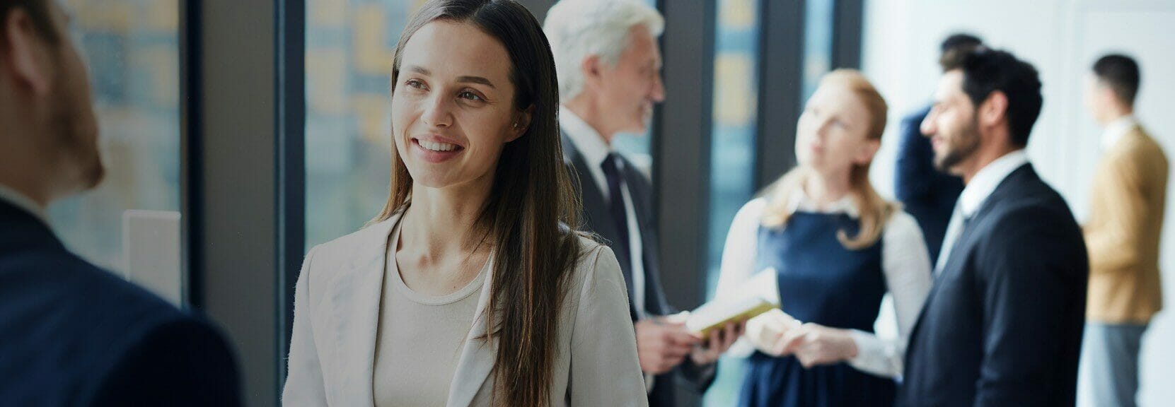 A business professional in a tan business casual outfit smiles at a man in a suit while other business professionals mingle in the background.