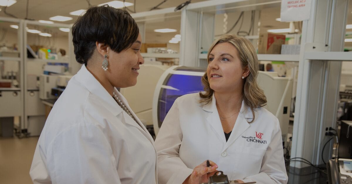 Two medical laboratory science professionals wearing University of Cincinnati lab coats discussing test results in a modern clinical laboratory.