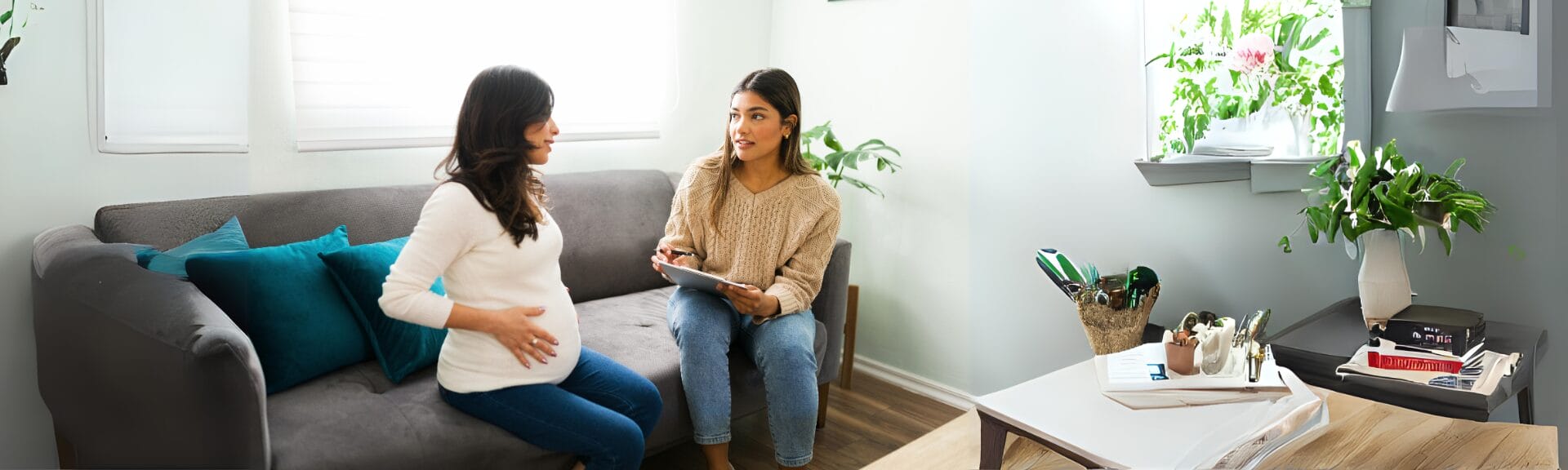 An image of a nurse midwife speaking with a pregnant woman while both are sitting on a grey sofa.