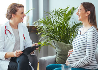 An image of a nurse midwife speaking with a pregnant woman while both are sitting on a grey sofa.