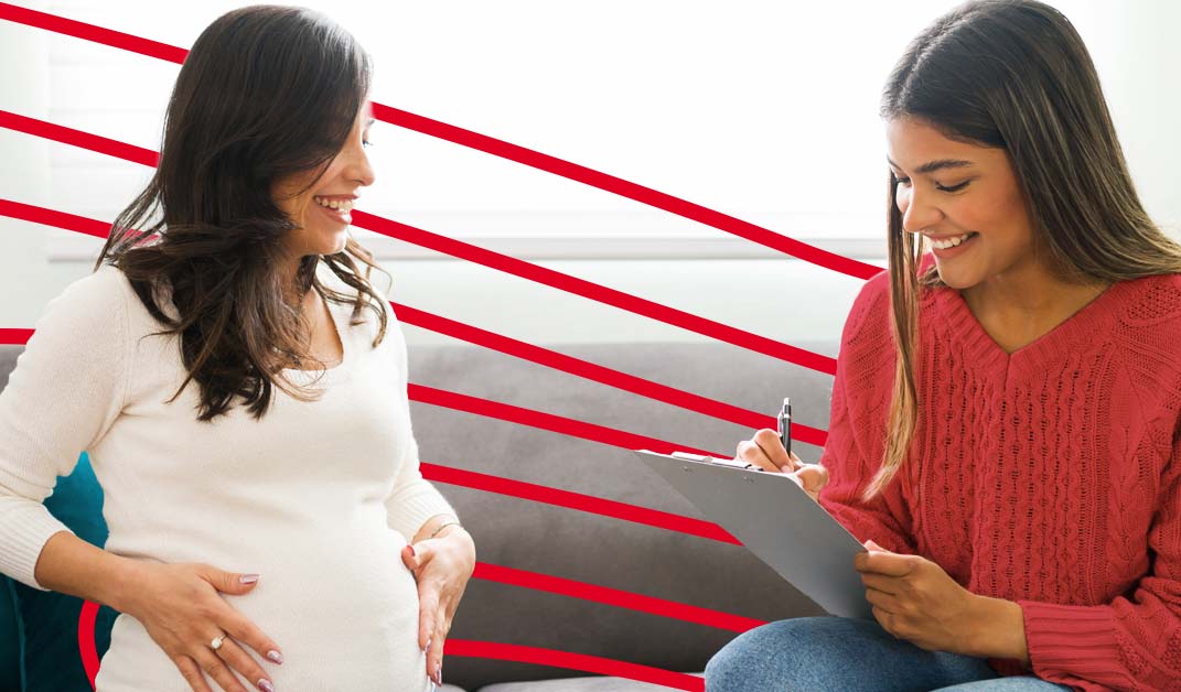 An image of a nurse midwife speaking with a pregnant woman while both are sitting on a grey sofa.