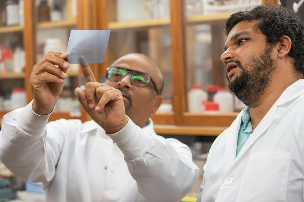 Two pharmaceutical science researchers in lab coats examining a film sheet or test result in a laboratory setting.