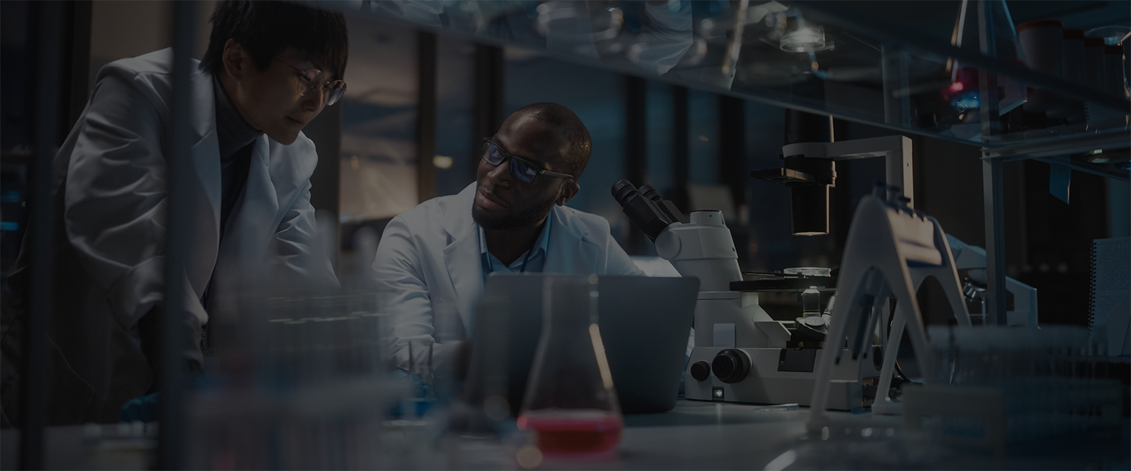 Researchers in a dimly lit laboratory discussing data beside microscopes and lab instruments, symbolizing health outcomes and pharmacoeconomics research.