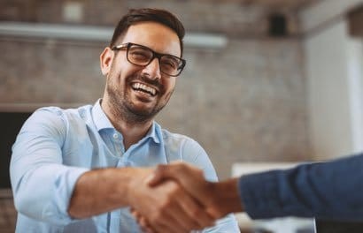 A man wearing a blue collared shirt and black-framed glasses shakes someone's hand. The other person's full body is out of frame and only their forearm is visible.