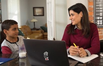 A mom looks at her son while she is working on a laptop that has a University of Cincinnati C-Paw logo sticker on the center.