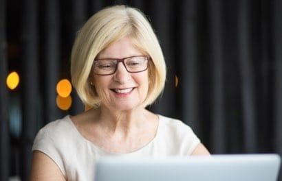 A blonde woman wearing black-framed glasses and a white T-shirt smiles while working on a laptop.