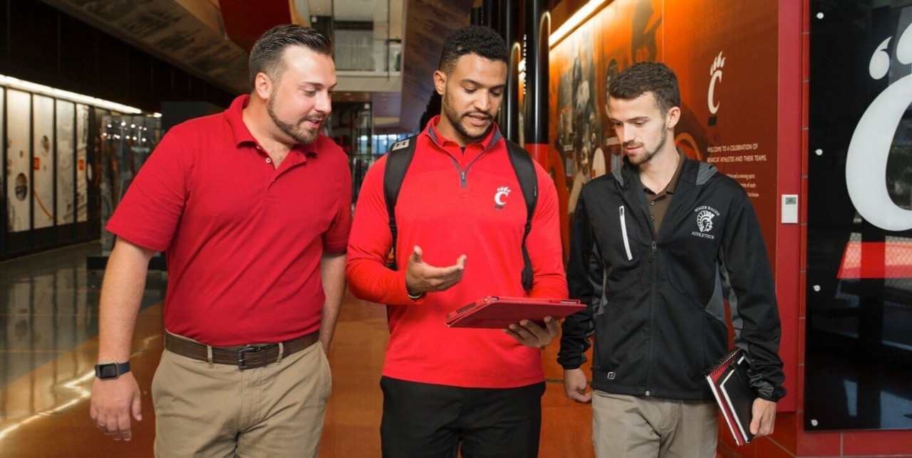 Three sport admission male students walking and looking at a tablet that is held by the man in the middle.