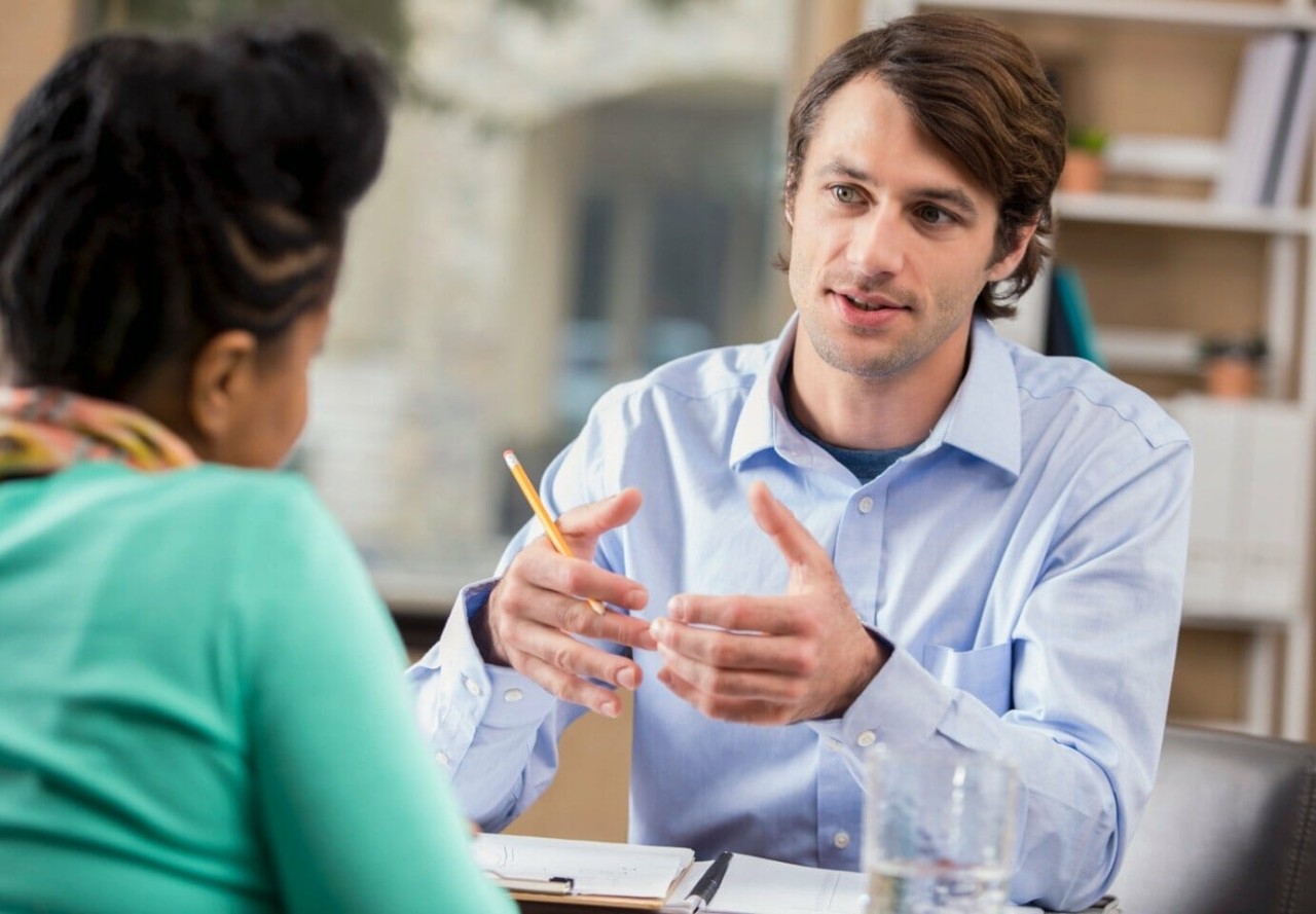 A male advisor in a light blue, collared shirt holding a pencil speaks with a woman wearing a teal sweater. Both are seated at a desk.