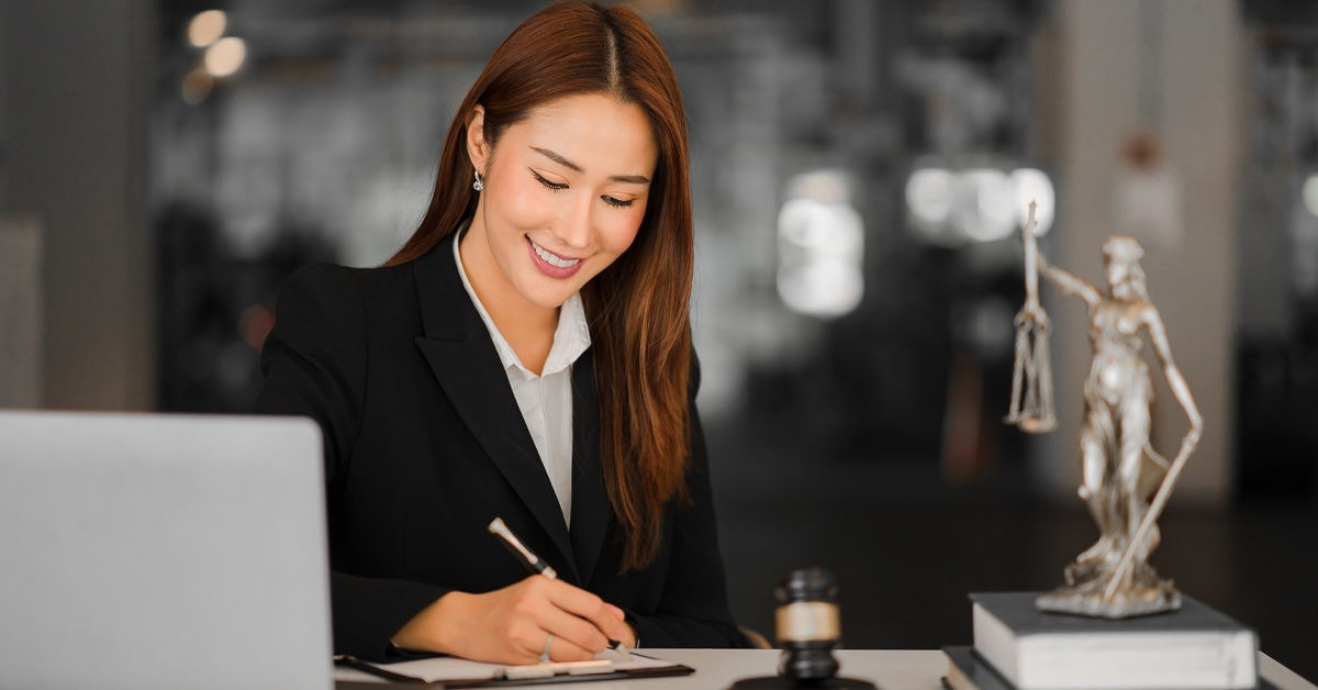 A woman wearing a black blazer sits writing at a desk in a law office. On her desk is a small bronze statue of Lady Justice, as well as a judge's gavel. 