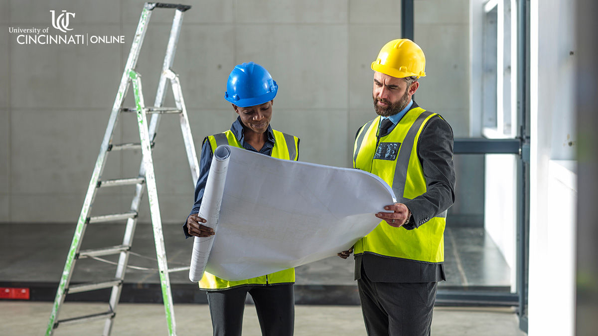 Two city planners stand and look at city plans on paper. One planner wears a blue hardhat and the other wears a yellow hardhat. 