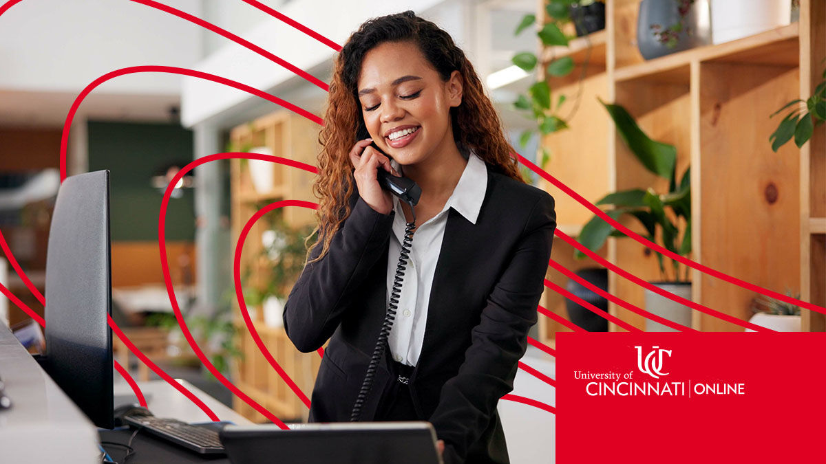 Female hospitality worker in white shirt and black blazer smiles as she talks on the phone at a hotel front desk.