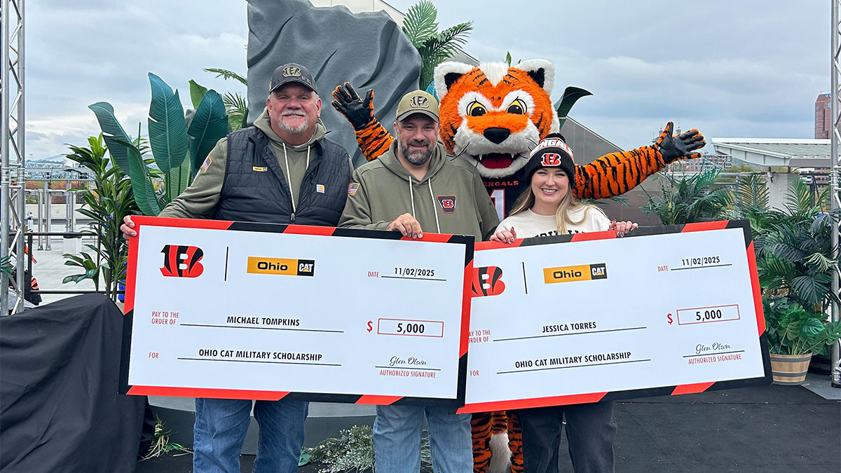 Two men and one woman stand onstage with the Bengals tiger mascot, all wearing Cincinnati Bengals attire. They hold two oversized checks that display two $5000 military scholarships.