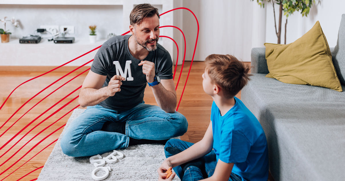 A male speech language pathologist in a dark grey t shirt and jeans sits on the floor and speaks with a young boy. The man holds up a plastic letter M, and other letters are on the carpet between them.