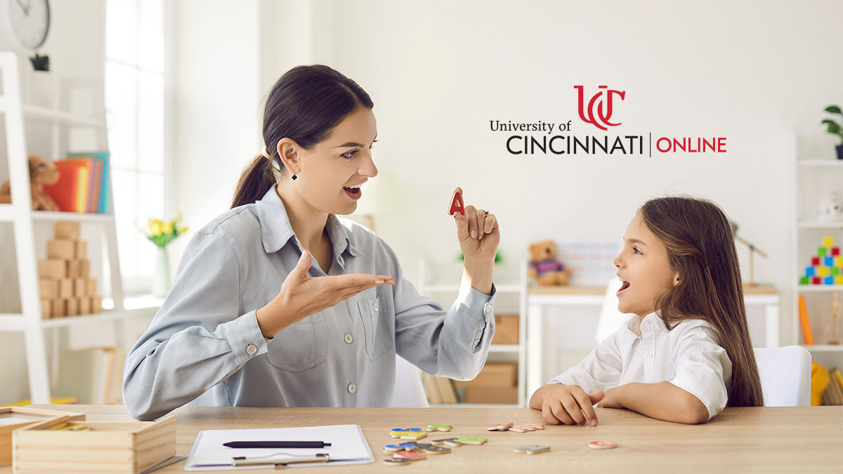 A female speech-language pathologist in a light blue button-down shirt gestures to a red letter A. She shows it to a young girl with long brown hair wearing a white shirt. They sit in an SLP office.