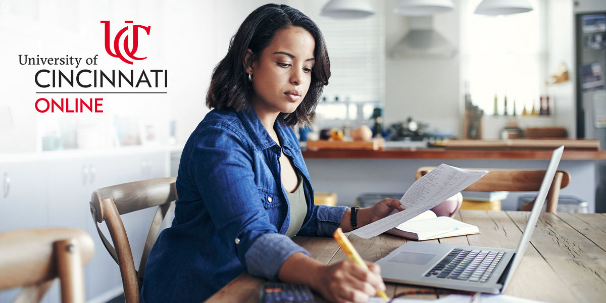 A black woman with short straight hair sits at a kitchen table wearing a jean jacket and olive green tank top. She sits in front of her laptop with paper in one hand and a pencil in the other.