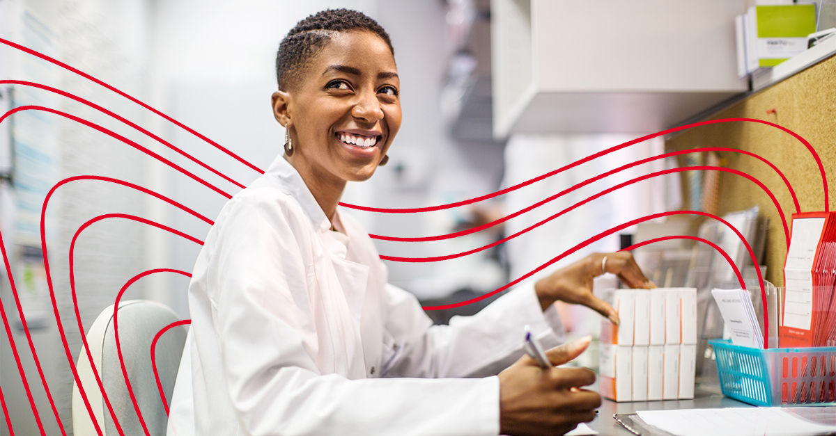 A black woman with buzzed dark hair wears a lab coat and sits at a medical office desk. She holds a pencil and smiles to her right.