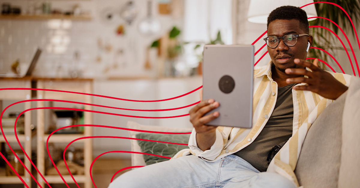 A male college student sits on a couch at home. He is holding up a gray tablet and talking to the screen for an online therapy appointment.