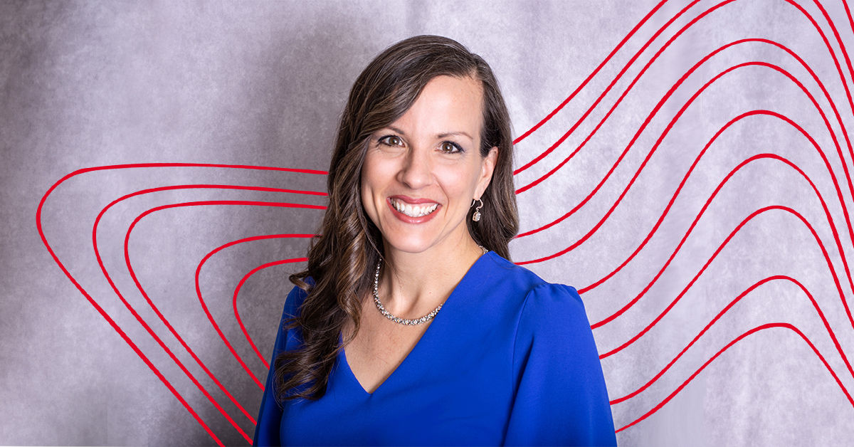 A woman with long brown hair wears a long sleeve blue blouse and smiles in front of a gray background.