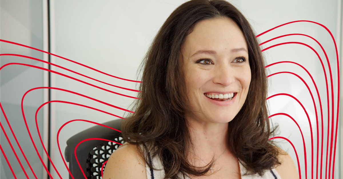 A woman with shoulder length brown hair sits in front of a white and red background and smiles at an offscreen interviewer.