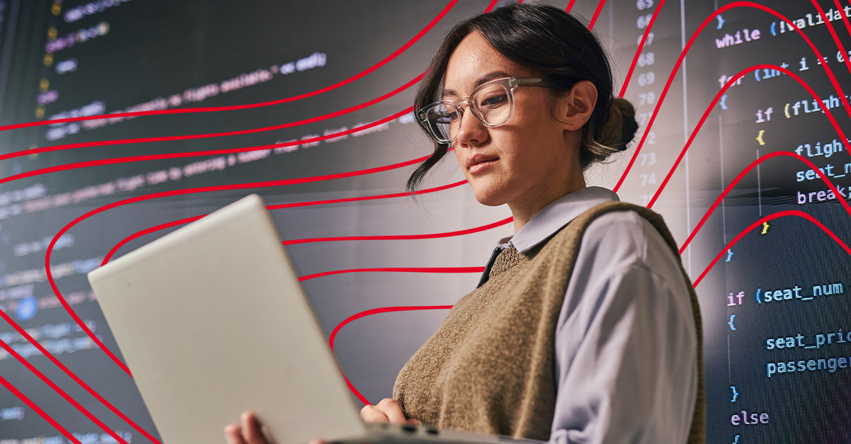 A woman wearing glasses and a tan sweater vest stands in front of a large screen projection of code. She holds a white laptop and scrolls on it.