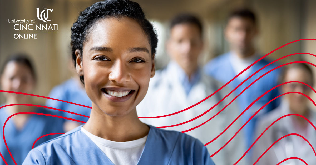 A nurse in blue scrubs smiles into the camera. Four other healh care professionals stand in the background and also look at the camera.
