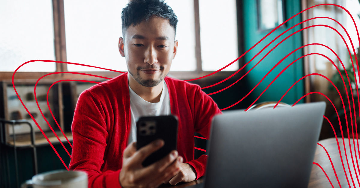A man in a white t-shirt and red cardigan sits on a couch in front of a laptop. He is in front of a bright window and holds a black phone in his hand.