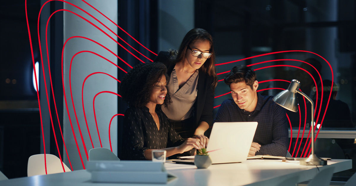 A woman stands and a man and woman sit at a desk in dimly lit office. They all look at silver laptop lit by a desk lap.
