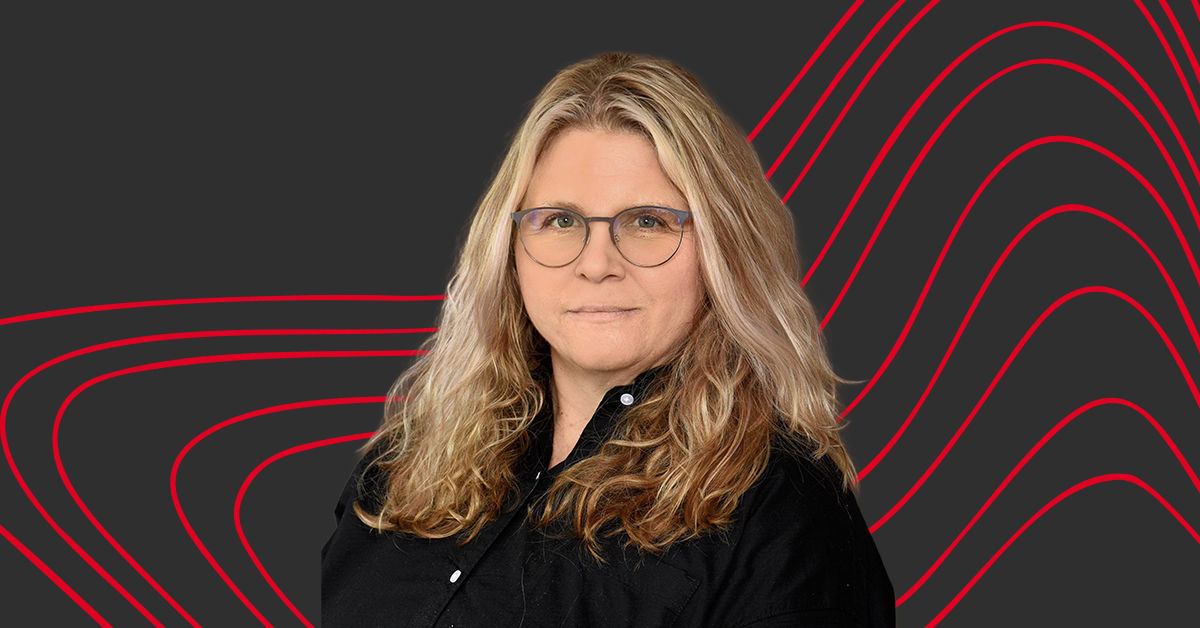 Amanda Feltner, a master of legal studies graduate, stands in front of a grey and red striped background. She wears a black button down shirt and glasses and has blonde hair.