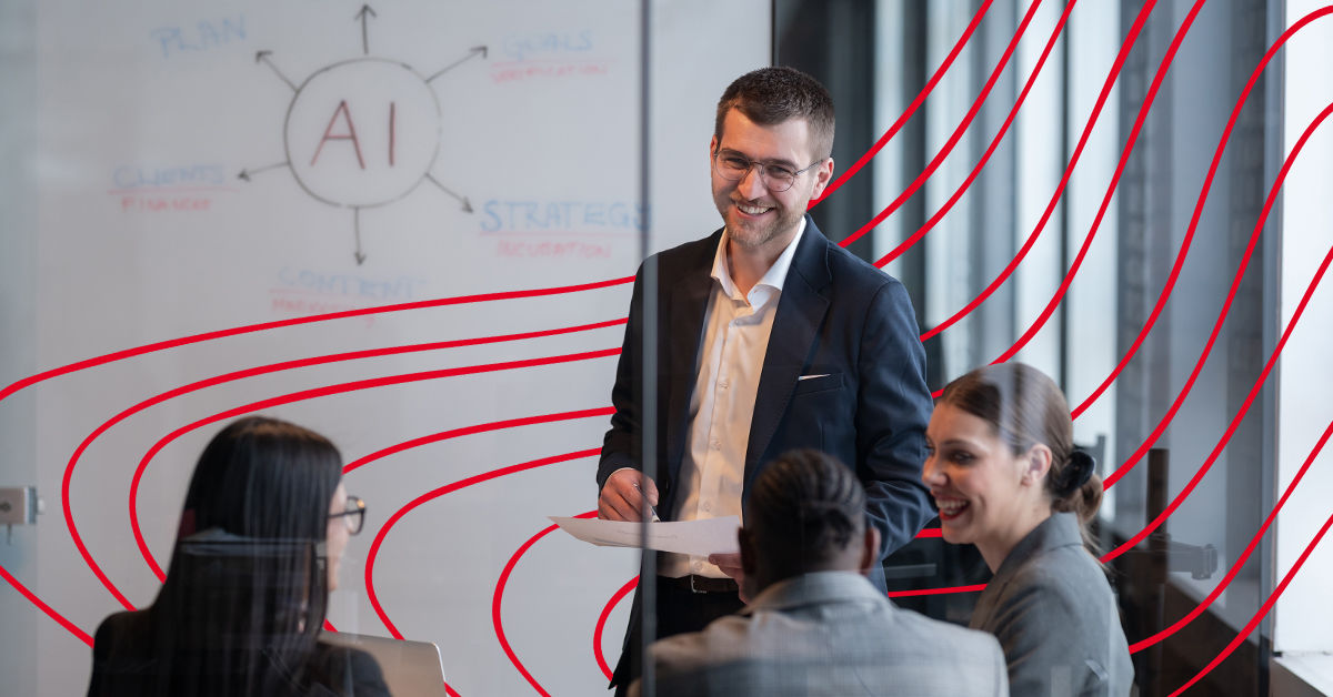 A man stands in a conference room in front of a whiteboard. On the whiteboard is a diagram with AI written in the center. Two women and a man in professional clothing sit at a table and listen to the man presenting.