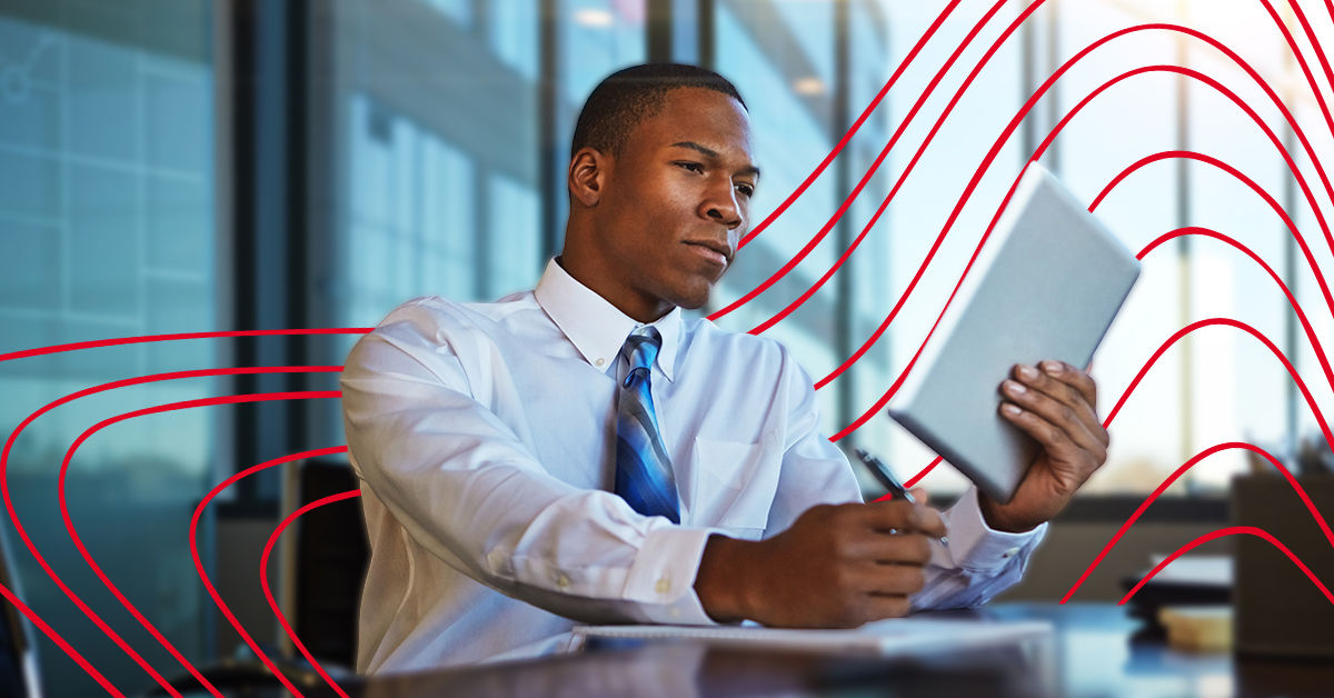 A black man wearing a white button up shirt and blue tie sits at a desk and holds a tablet. He writes on paper on the desk. In the background is a brightly lit office space and window.
