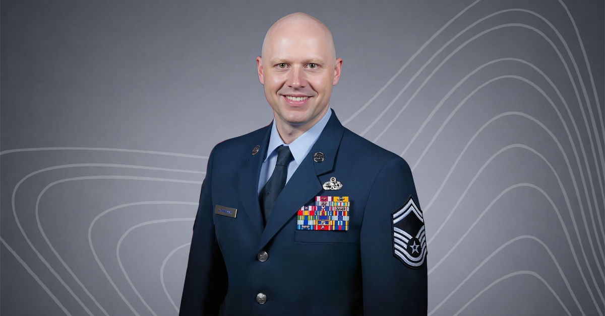 A man in the US Armed Forces wears a navy military uniform and smiles at the viewer. He is bald, and beneath his uniform jacket is a light blue collared shirt and a navy tie.