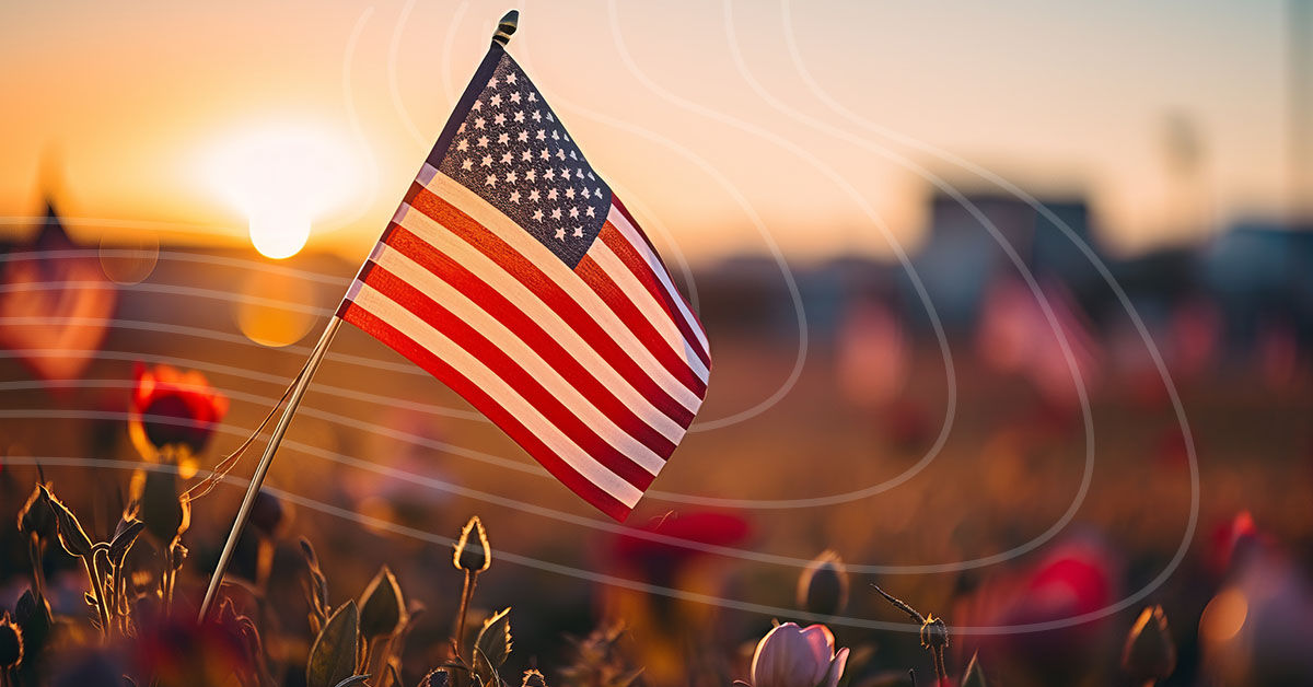 A close-up of the American flag surrounded by flowers with the warm glow of a sunset behind it.