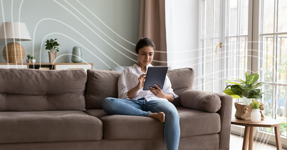 A woman with dark hair pulled back into a bun sits on a drown couch in her living room. She wears a casual white blouse and blue jeans. She holds a tablet and looks at the screen as she receives mental health support.