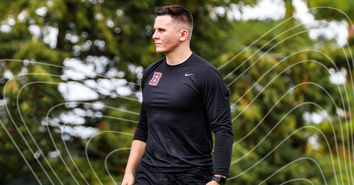 A man in a black long sleeve athletic shirt stands on an outdoor football field in front of a forested background. He has short brown buzzed hair, and he looks to his right with determination.
