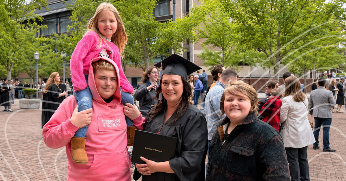 UC online graduate Emily Schnicke stands outside on UC's campus in a black graduation robe and cap. She stands with her three children; two are boys and wear a black pullover and a pink pullover, while the third daughter sits is on one of the son's shoulders in a pink sweatshirt.