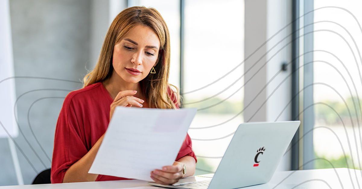 A woman sits at a desk in a brightly lit office. She sits in front of a laptop with a UC logo and looks at a piece of paper. She has long blonde hair and wears a red cardigan.