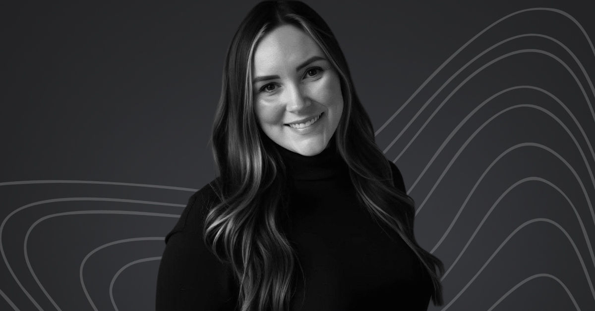 A woman in a black turtleneck with long brown hair smiles. She stands against a black background with grey swirls. 