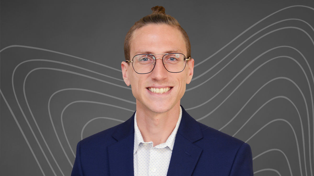 Online MBA graduate Justin Lincoln smiles and wears a dark navy blazer over a white collared shirt. He weares glasses and has light hair worn in a high bun.