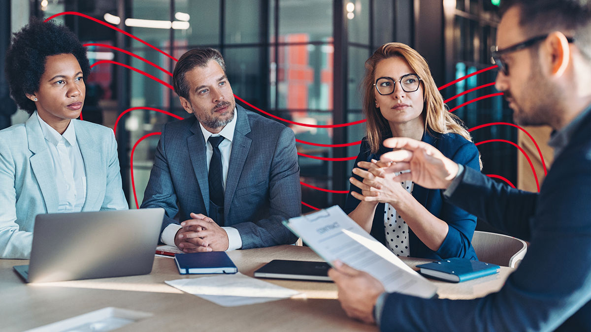 In a office meeting room, two men and two women sit at a table. One man in thick black glasses holds a clipboard with notes and presents information to his colleagues, who look at him in rapt attention. Computers and notebooks sit on the table. 