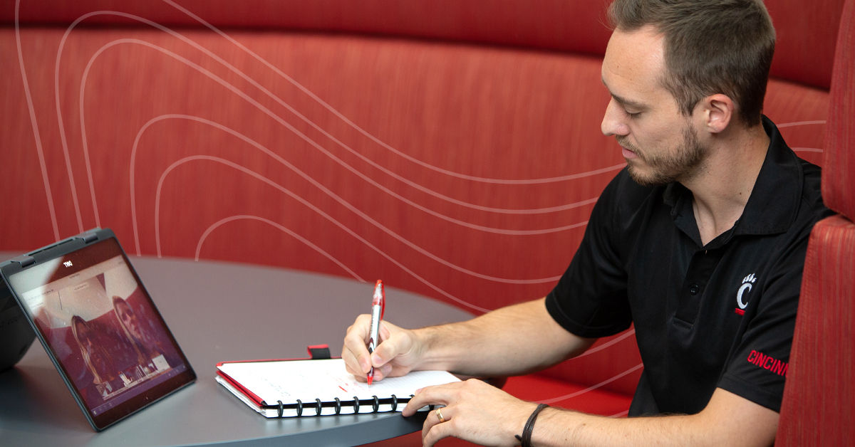 A white man with short brown hair sits at a gray table and writes in a notebook. He’s wearing a University of Cincinnati black polo shirt and watching an online lecture on a tablet in front of him.
